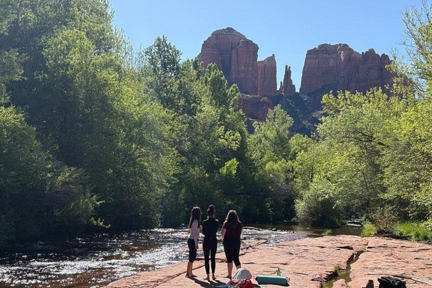 A river with people standing on a rocky outcrop, surrounded by dense green trees and red rock formations in the background. 2