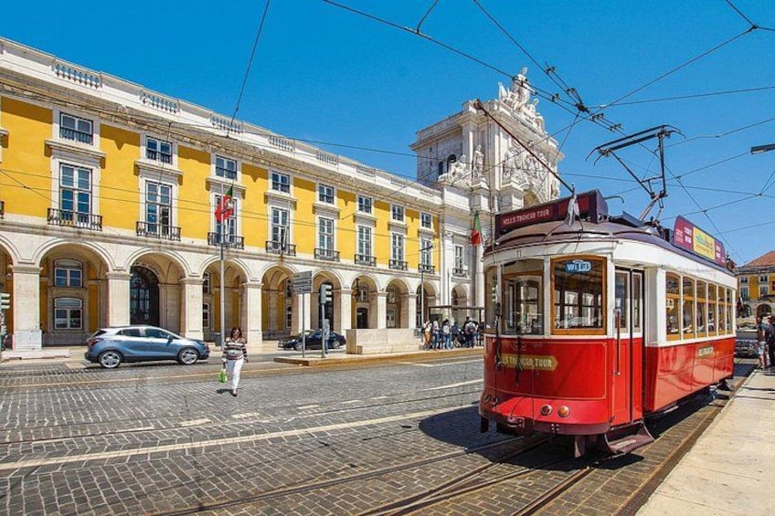 Un tramway rouge sur les rails, un bâtiment jaune avec des fenêtres en arc de cercle et une statue au sommet. 1