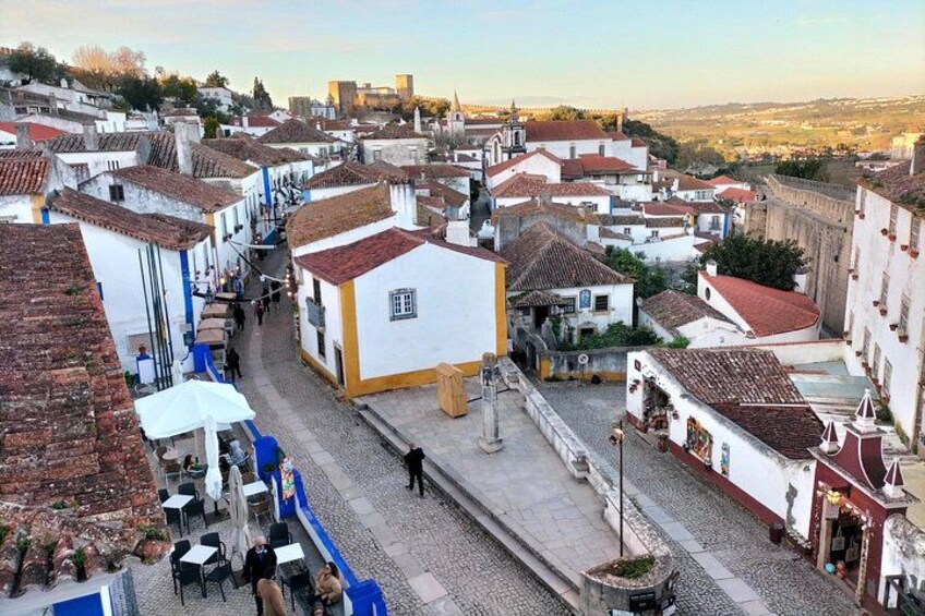 A cobblestone street lined with white buildings with red-tiled roofs, a person walking, and outdoor seating areas. 4