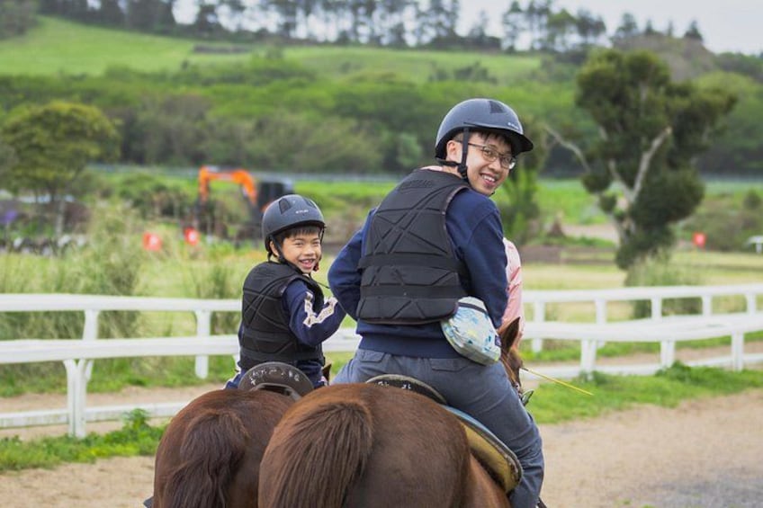 Two people riding horses on a dirt path with a white fence and green fields in the background. 2