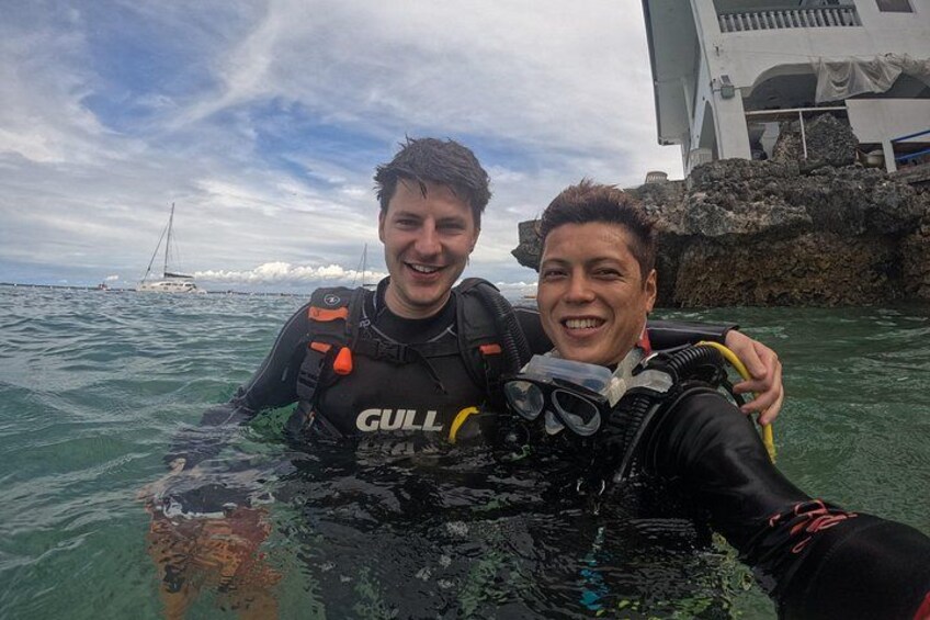 Two people in scuba diving gear with a rocky shoreline and a sailboat in the background. 3