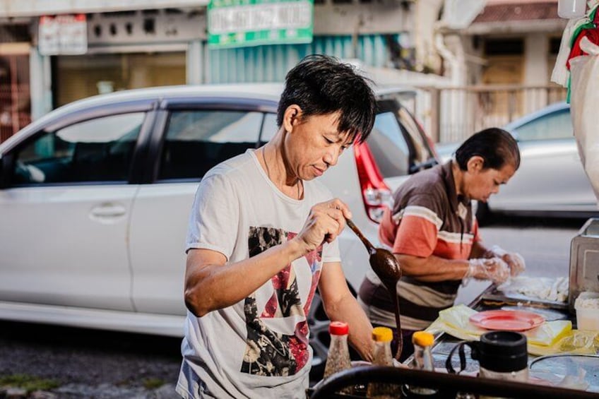 Two people cooking outdoors with a car and buildings in the background. 2
