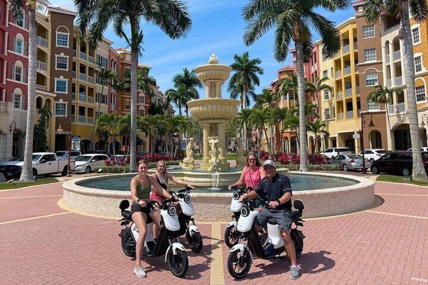 A couple on scooters in a plaza with a fountain and palm trees. 1