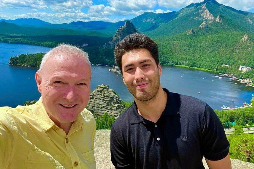 Dos personas frente a un paisaje montañoso con un lago y cielo despejado. 4