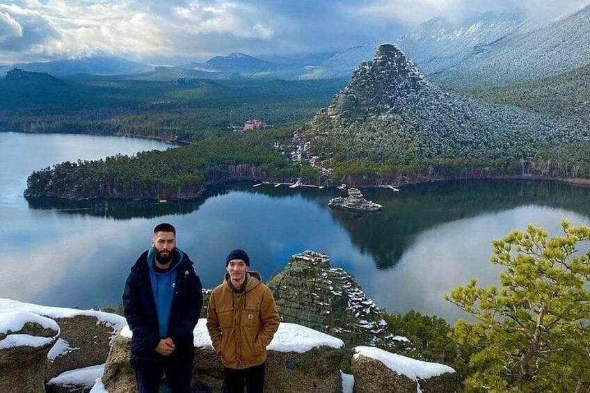 Dos personas de pie sobre un saliente rocoso, con un lago y montañas al fondo. 2