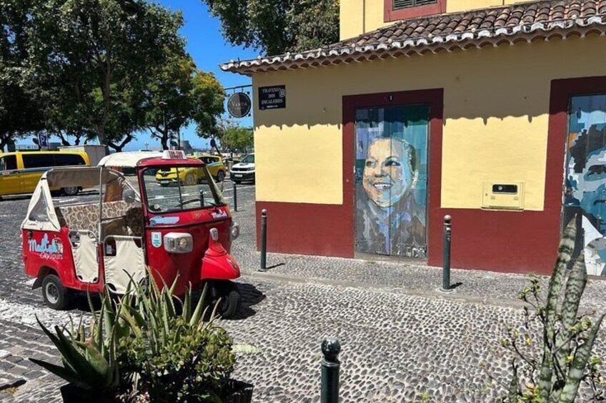 A red tuk-tuk parked in front of a building with a mural, a yellow taxi in the background, and a cobblestone street. 1