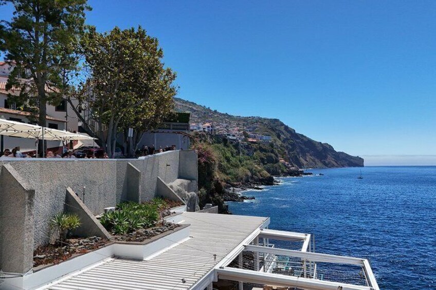 A coastal promenade with steps leading to a terrace overlooking the sea, a hillside with buildings, and a clear blue sky. 4