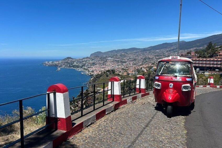 A red vehicle on a coastal road with a red and white guardrail, overlooking a coastal town and the ocean. 5