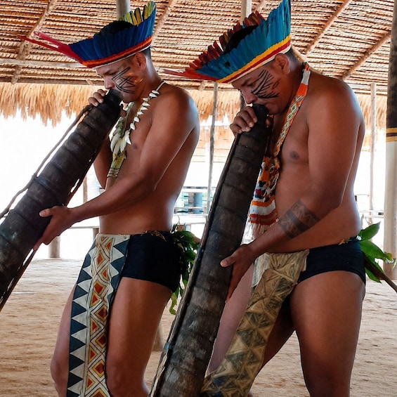 Two individuals in traditional attire holding large wooden objects, possibly ceremonial items, under a thatched roof structure. 4