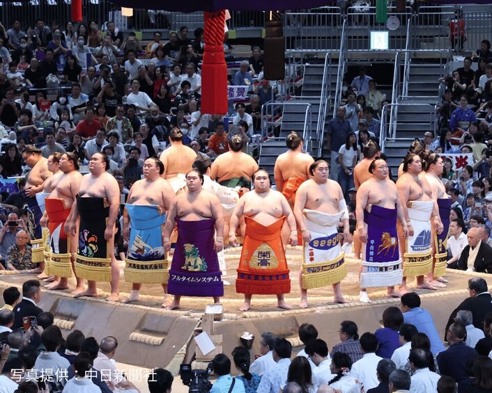 A group of sumo wrestlers standing in a line on a stage with spectators in the background. 1