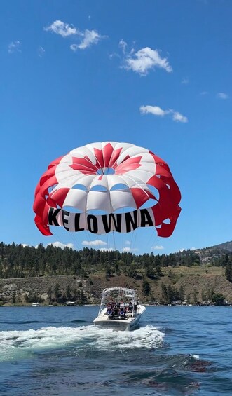 A parasail with the word 'KELOWNA' over a body of water with a boat and a forested hillside in the background. 1