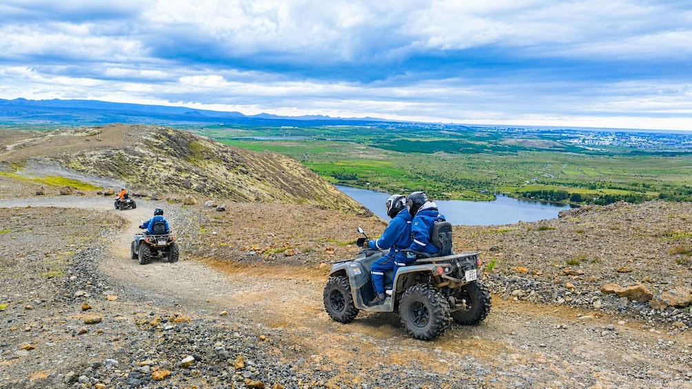 Three people on ATVs on a rocky path with a lake and mountains in the background. 1