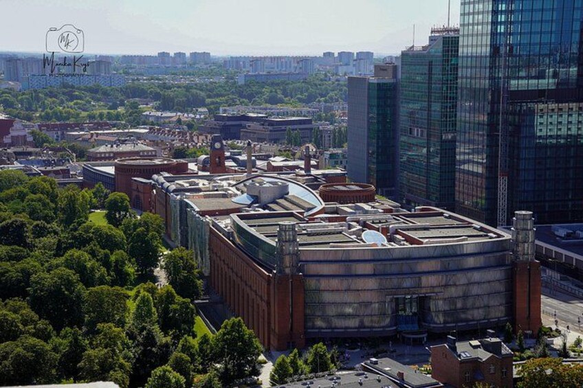 A cityscape with a large industrial building, modern skyscrapers, and surrounding greenery. 4