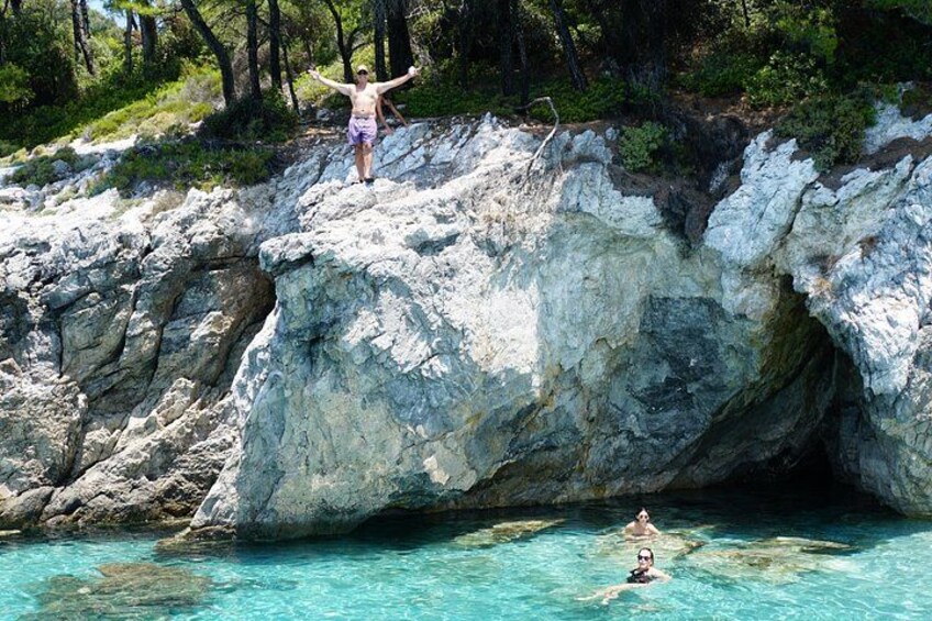 A person standing on a rocky cliff with arms outstretched, another person swimming in clear blue water below, and a forested area in the background. 2