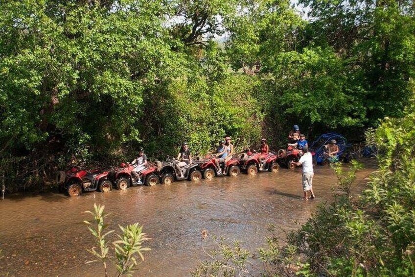 A group of people on ATVs crossing a shallow water body surrounded by dense greenery. 3