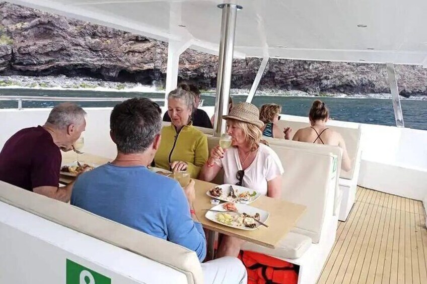 Un groupe de personnes est réuni autour d’un repas sur un bateau, avec des falaises rocheuses en arrière-plan. 5