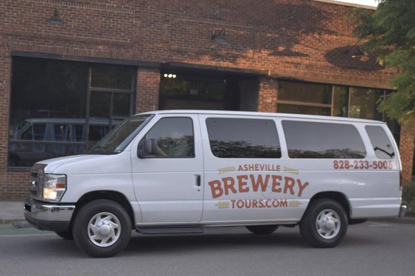 Una camioneta blanca estacionada frente a un edificio de ladrillo con las palabras 'Asheville Brewery Tours' y un número de teléfono en la carrocería. 2