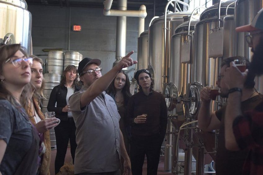 Un grupo de personas en una cervecería, observando y señalando el equipo de elaboración. 4