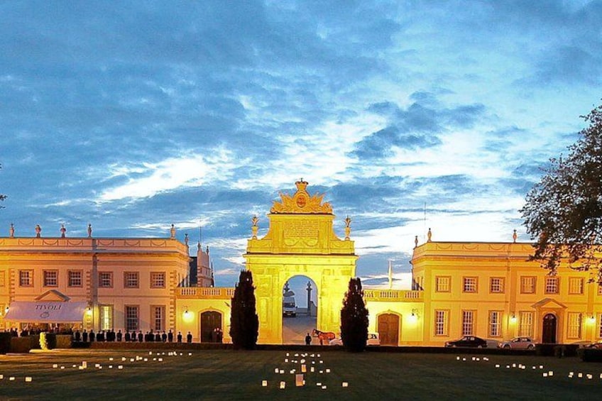 A grand illuminated archway with a statue on top, flanked by two buildings with classical architecture, under a twilight sky. 4