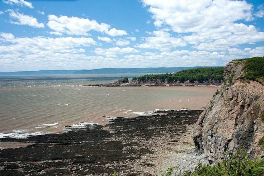 Eine Küstenlandschaft mit felsigem Ufer, einem Sandstrand und einer entfernten Berglandschaft unter einem teilweise bewölkten Himmel. 1