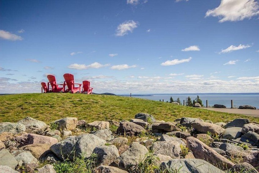 Red Adirondack chairs on a grassy hill with a rocky foreground and a body of water in the distance. 3
