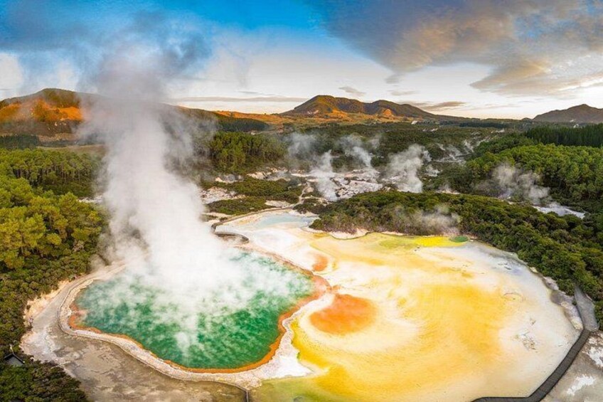 Un área geotérmica con respiraderos de vapor, aguas termales de color verde y amarillo, y un paisaje boscoso. 1