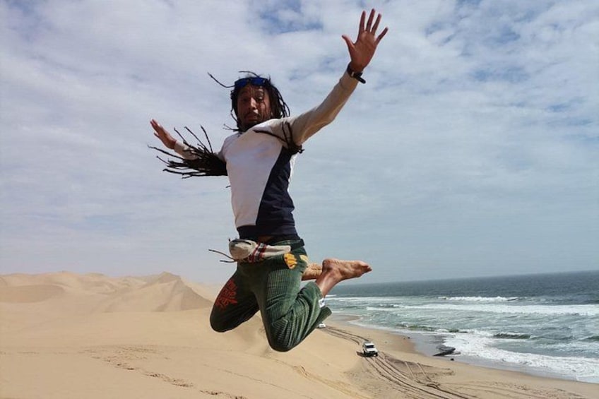 Una persona salta en una playa con dunas de arena y el océano al fondo. 5