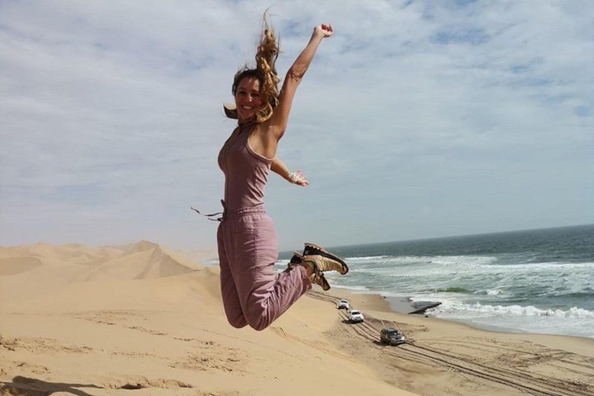 Una persona saltando en una playa con dunas de arena y el océano al fondo. 2
