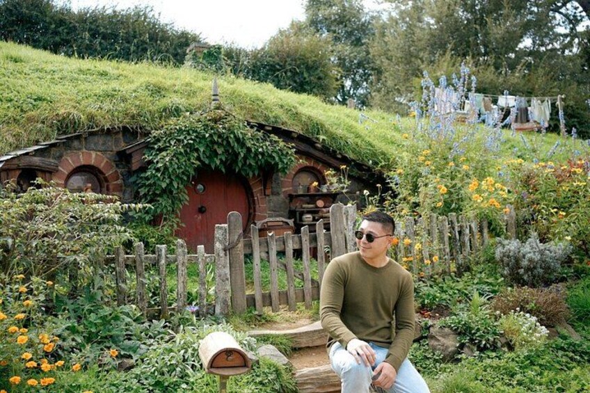 Eine Person sitzt vor einem hausähnlichen Bau im Hobbit-Stil mit einem Strohdach, umgeben von einem Garten mit verschiedenen Pflanzen und Blumen. 4