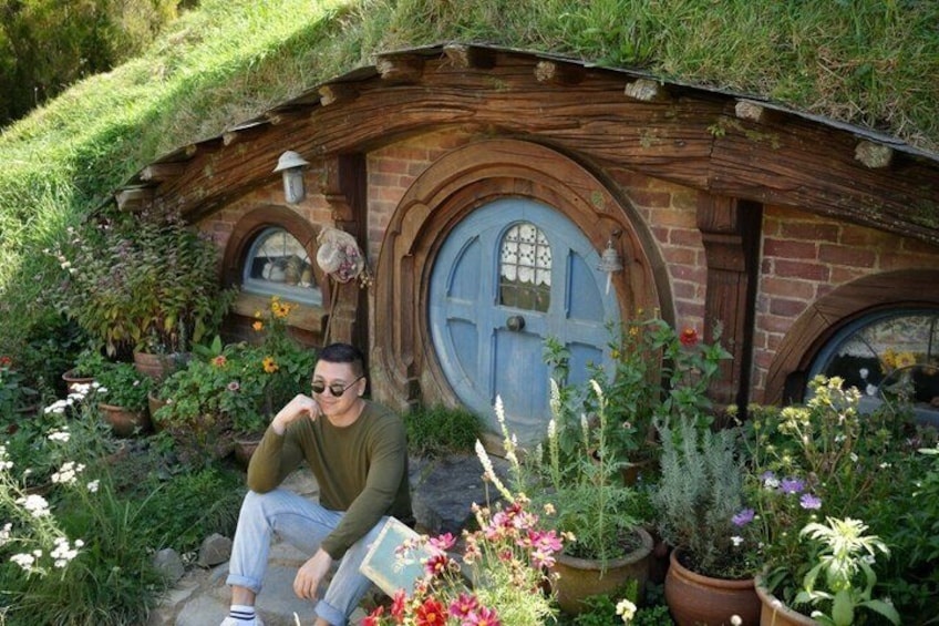Et hobbithuls-lignende hus med stråtækt tag, en blå dør og en have med forskellige krukker og blomster. 1
