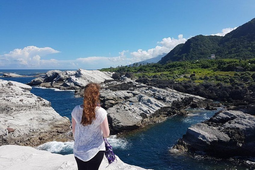 A person standing on a rocky shore overlooking a river, with mountains and greenery in the background. 1
