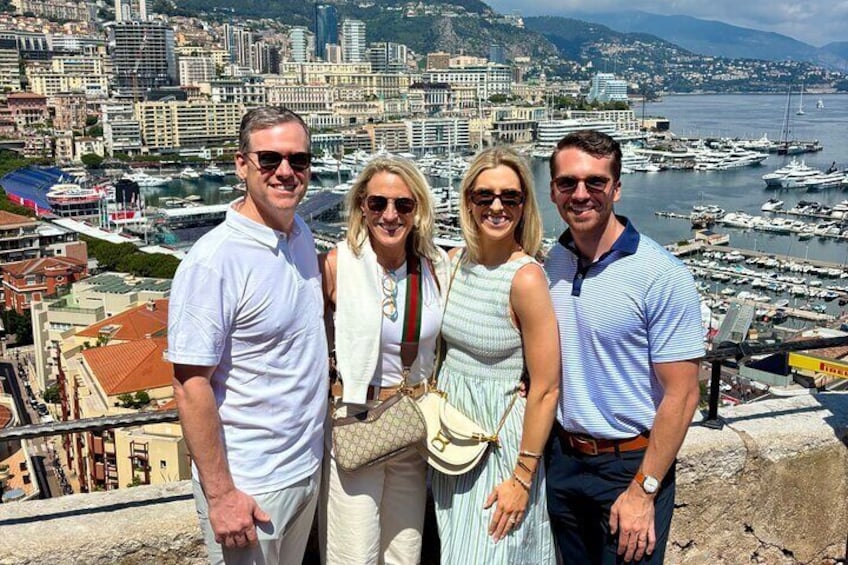 A group of people standing on a cliff overlooking a marina with boats and a cityscape in the background. 1