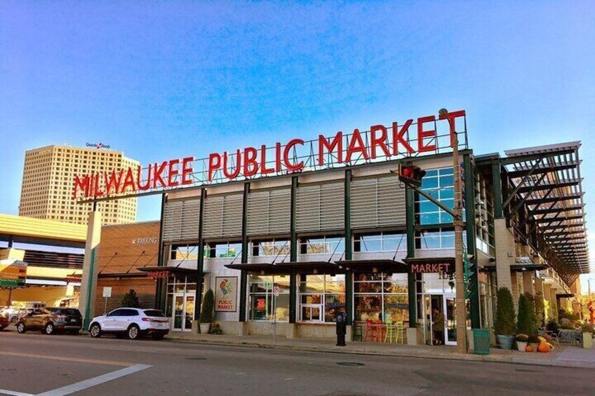 The exterior of the Milwaukee Public Market with a large sign, storefronts, and a parking lot. 4