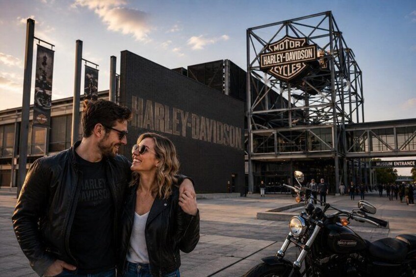 A couple standing in front of a Harley-Davidson museum entrance with a motorcycle parked nearby. 1
