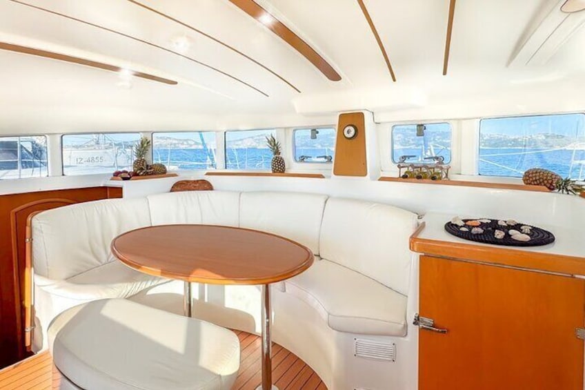 A boat interior with a white cushioned seating area, a wooden table, and a small kitchen counter with a sink. 3