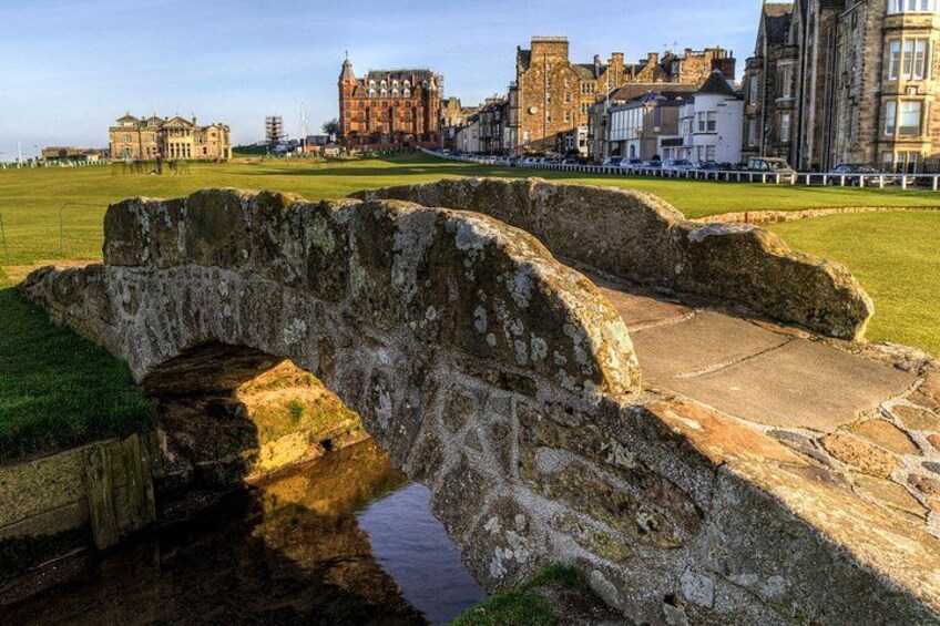 A stone bridge over a small stream with a grassy field and historic buildings in the background. 1