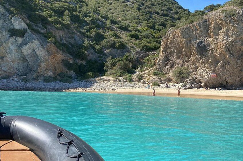 A clear turquoise sea, a sandy beach, rocky cliffs, and a boat with a black inflatable dinghy. 1