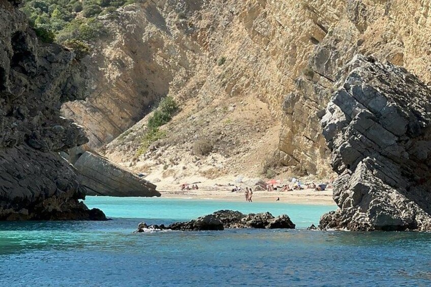A coastal landscape with rocky cliffs, clear turquoise water, and a sandy beach with people and umbrellas. 5