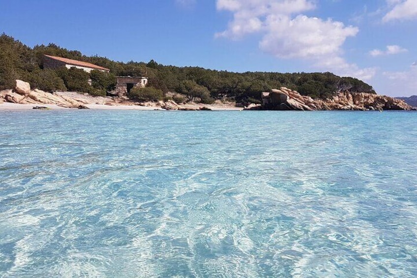 A clear coastal area with a sandy beach, rocky outcrops, and a few buildings on the hillside. 1