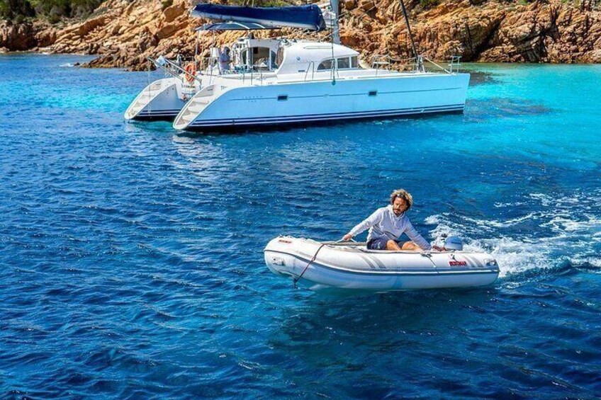 Une personne à bord d’un petit bateau gonflable sur une eau d’un bleu limpide, un bateau plus grand en arrière-plan et des falaises rocheuses. 2