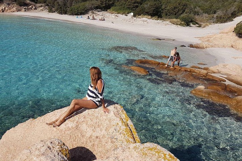 Een vrouw zittend op een rots aan een heldere blauwe zee, een zandstrand en rotsformaties in de verte. 5