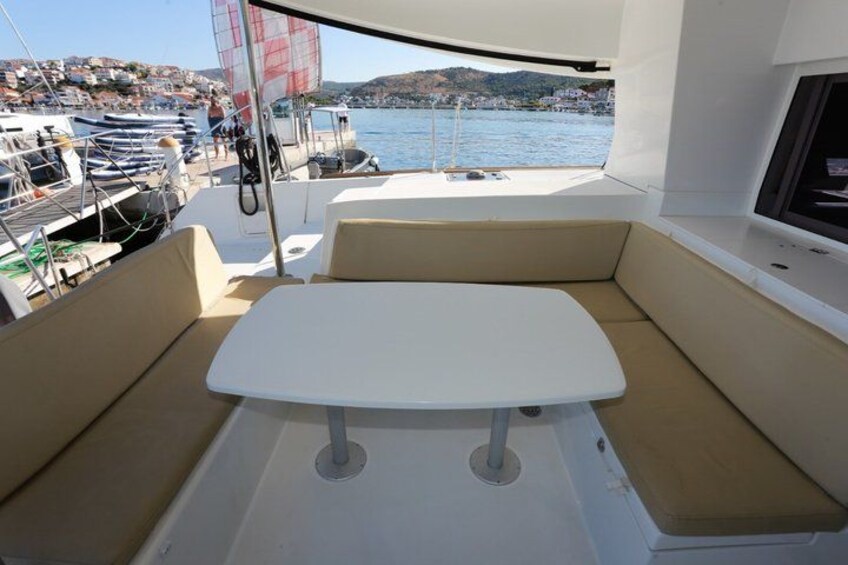 A boat's interior with a table and cushioned seating, a sail, and a view of a marina and hills in the background. 3