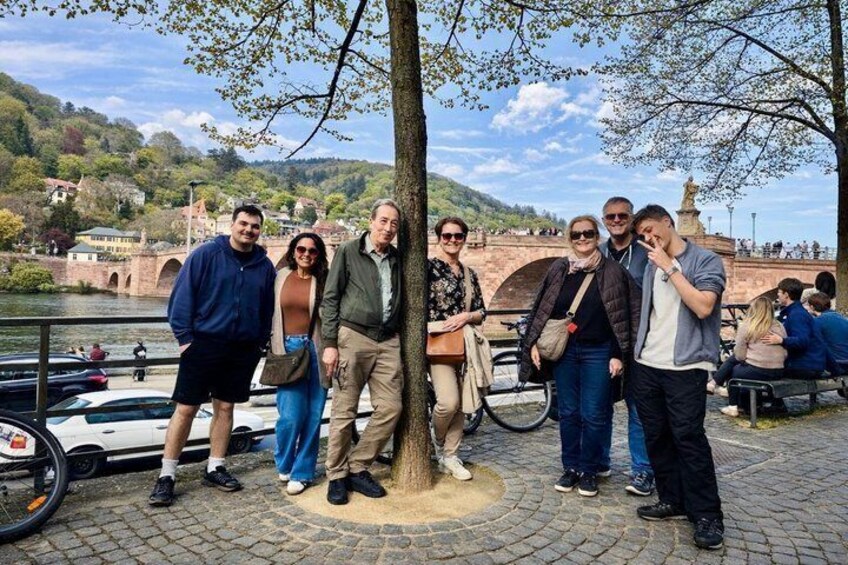 A group of people standing by a river with a bridge and hills in the background. 4