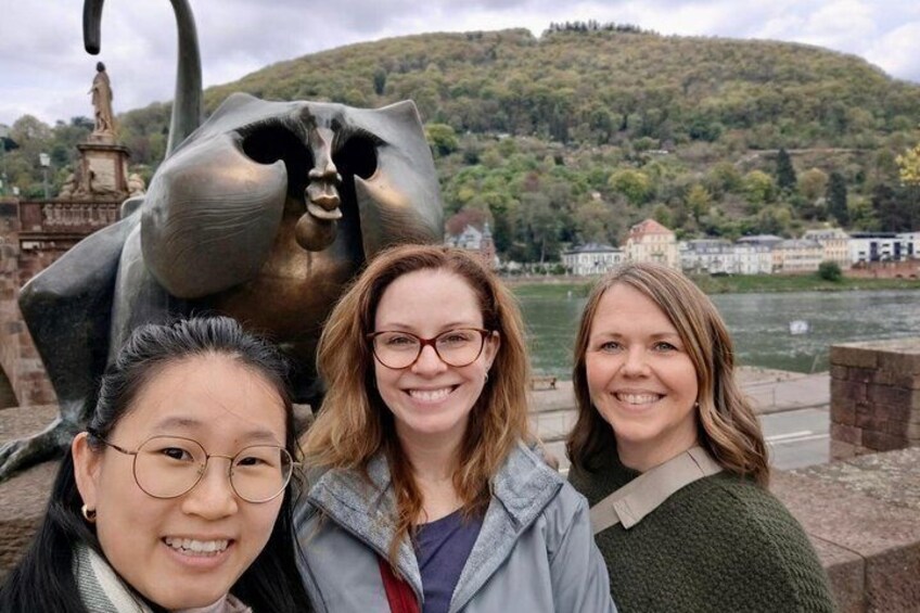 Three people in front of a large sculpture and a river with a hill in the background. 1