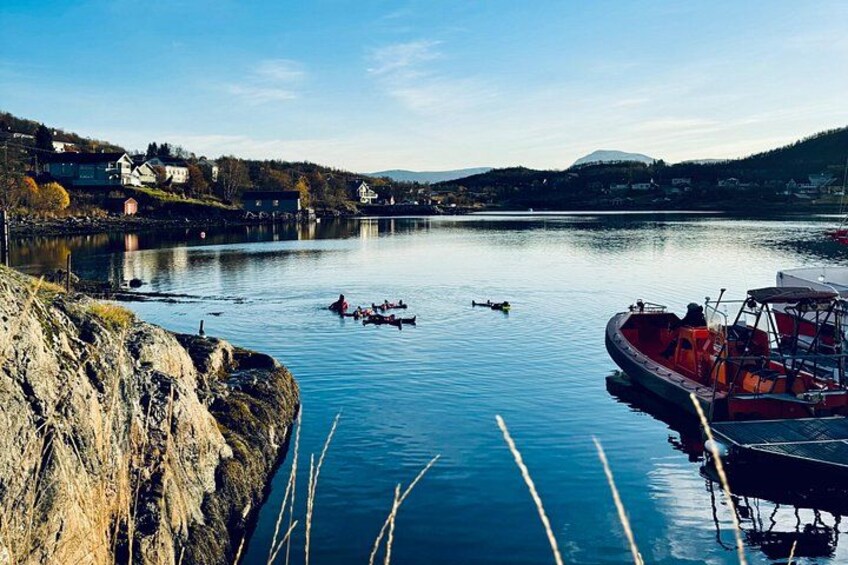 En rolig flod med en rød båd fortøjet til højre, en gruppe mennesker i en mindre båd i midten, og huse på venstre bred. 2