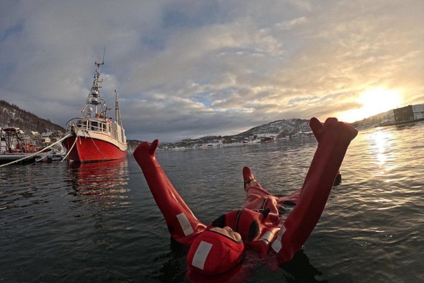 Une personne portant un gilet de sauvetage rouge flotte sur une eau calme, et un bateau rouge est visible à l'arrière-plan. 2