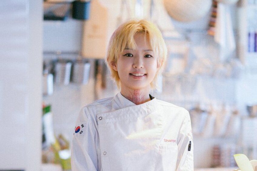 Une personne vêtue d’un uniforme de chef blanc, orné d’un logo sur la poitrine gauche, se tient dans une cuisine aux armoires blanches, avec divers ustensiles de cuisine en arrière-plan. 2
