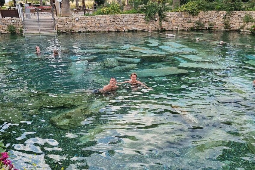 Una piscina con agua cristalina, personas nadando y un muro de piedra al fondo. 1