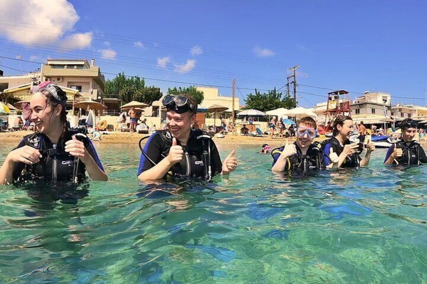People in clear water, wearing diving gear, with buildings and umbrellas in the background. 1