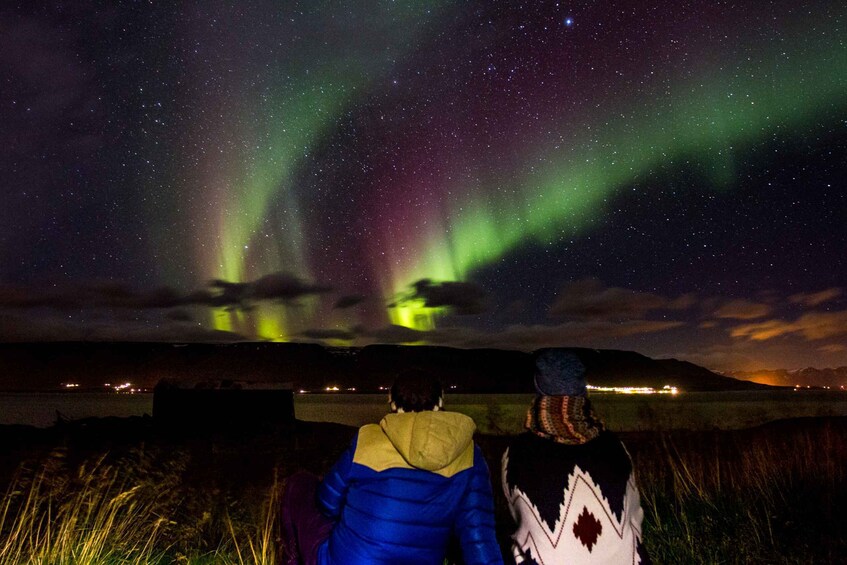 Two people observing the Northern Lights over a body of water with mountains in the background. 2